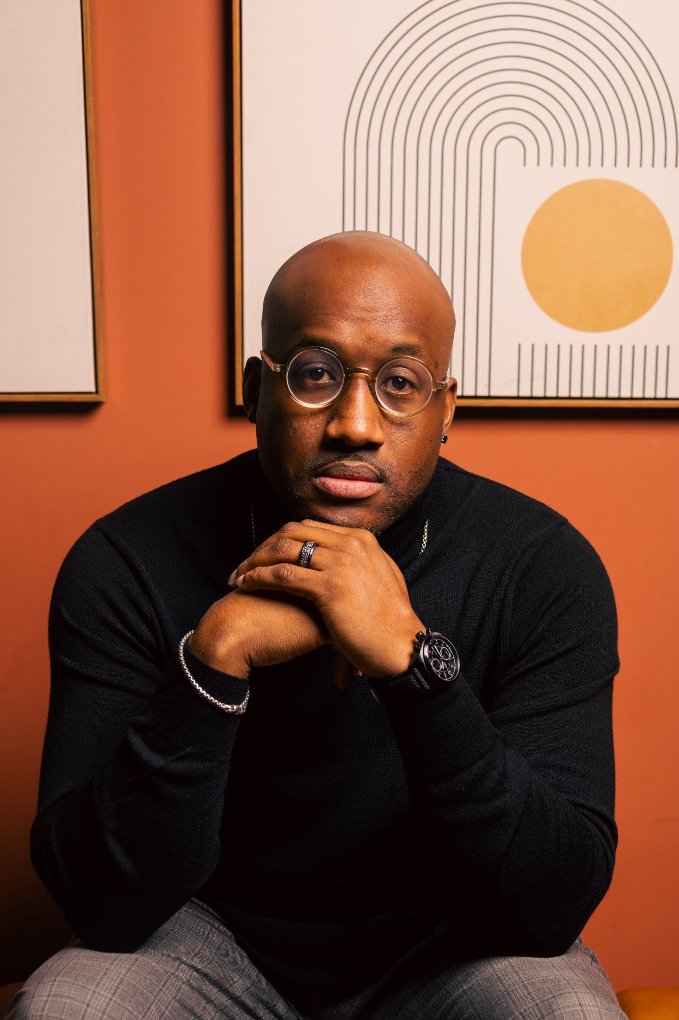 Lonnell Williams close-up portrait, black turtleneck, hands clasped, direct gaze against orange wall